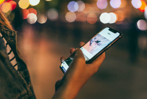 Person holding a mobile phone in front of blurred city lights in the evening.